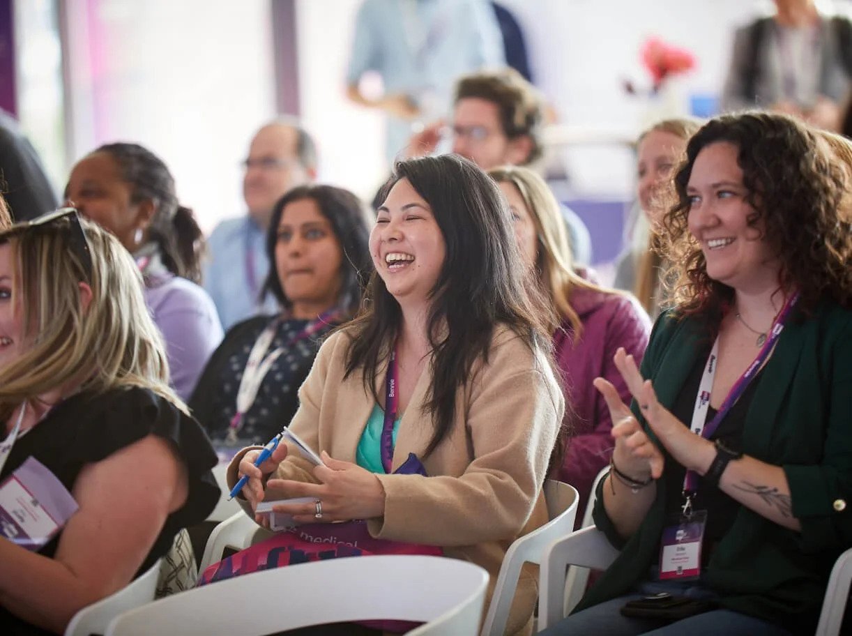 A group of people seated indoors, smiling and clapping, appearing engaged at an event or conference.